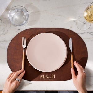 May include: A dark brown oval placemat featuring the text "MAUN GENUINE CUISINE" in gold lettering. A light pink plate sits in the center, flanked by a silver fork and knife with wooden handles. A glass of water and a wine glass are also present.