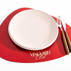 May include: A dining table setting featuring a white plate, a fork, and a knife with wooden handles. The plate rests on a red placemat with the text "VINEYARD Club" in gold. The setting is on a white background.