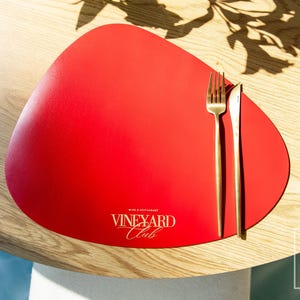 Könnte beinhalten: Ein roter, tropfenförmiger Platzdeckchen mit einem goldenen Gabel- und Messerset auf einem Holztisch. Auf dem Platzdeckchen steht "Vineyard Club". Der Tisch ist rund und hat eine helle Holzmaserung.