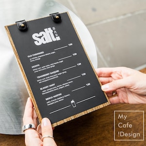 Puede incluir: Una pizarra de menú negra con el texto "salt bar" en blanco, que enumera cócteles. La pizarra está sostenida por manos, y el fondo incluye una mesa metálica y una superficie de madera. El menú enumera cócteles clásicos.