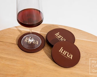 May include: A wine glass with red wine sits on a brown leather coaster. Two additional brown leather coasters with the gold text "luna restaurant" are next to the glass. The coasters are on a wooden surface.