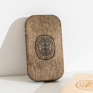 Può includere: Un portamenù rettangolare in legno con angoli arrotondati e una finitura tinta scura. La parte anteriore presenta un timbro nero circolare con il testo The Oriental Restaurant Premium Quality Food. Sono visibili due piccole cerniere metalliche color oro.