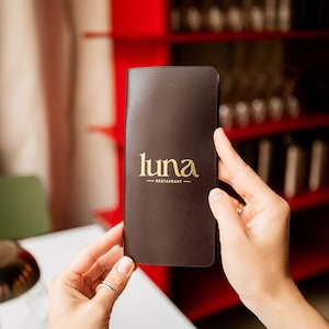 May include: A brown restaurant menu with the gold text "luna restaurant" is held by two hands. The menu is rectangular and appears to be leather-bound. The background includes a red shelf and a table.