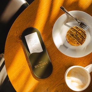 May include: A wooden table with a green leather menu holder that says "The Hungry" on it. There is a white plate with a pastry on it and a white coffee cup with a brown liquid inside.