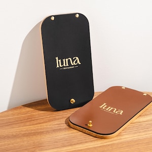 Może przedstawiać: Dwa prostokątne uchwyty na menu z zaokrąglonymi rogami. Jeden jest czarny, drugi brązowy. Oba mają słowo "luna" złotymi literami. Uchwyty znajdują się na drewnianym stole. Czarny uchwyt jest w pozycji pionowej, a brązowy leży.
