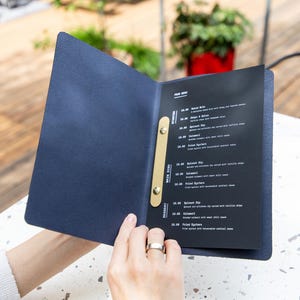 May include: A navy blue menu is open, revealing a black page with white text. The menu is held by a person. The menu has a gold-colored spine. The background is a wooden deck and a white table.