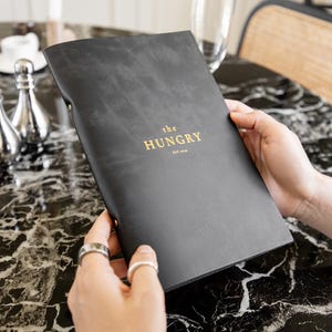 Könnte beinhalten: Ein schwarzes Menü mit goldfarbenem Schriftzug "the HUNGRY est. 2016" wird offen gehalten. Das Menü liegt auf einem schwarzen Marmortisch. Silberne Dekorationsartikel und eine weiße Kaffeetasse sind im Hintergrund.