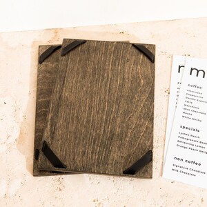 May include: Two rectangular dark brown stained wooden menu boards featuring black metal corner accents. Beside the boards is a white paper menu with black text listing coffee, specials, and non-coffee drinks, including items like Americano, Cappuccino, and Lychee Peach.