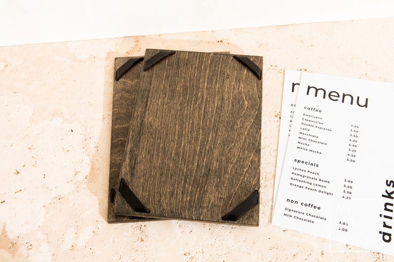 May include: Two rectangular dark brown stained wooden menu boards featuring black metal corner accents. Beside the boards is a white paper menu with black text listing coffee, specials, and non-coffee drinks, including items like Americano, Cappuccino, and Lychee Peach.