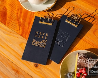 May include: Two navy blue menu clipboards with gold-colored clips and the text "WAVE CAFE" in gold. One clipboard has a gold wave design. A slice of cake with berries sits on a plate on a wooden table.
