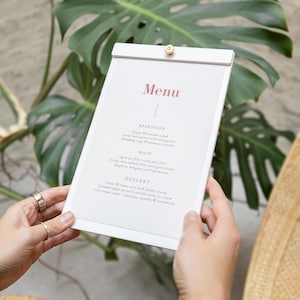 Puede incluir: Un menú blanco con la palabra «Menú» en fuente burdeos. El menú está sostenido por manos y tiene un cierre dorado en la parte superior. El menú enumera aperitivos, platos principales y postres. El fondo incluye hojas verdes y una superficie tejida.