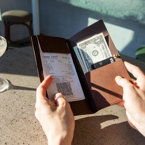 Op de afbeelding: Een bruine leren bonhouder met een bon en contant geld erin. De bon toont een totaal van 6,32. Een creditcard is ook zichtbaar. Een cocktail met een sinaasappelschijfje ligt op tafel.