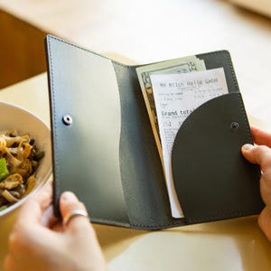 Puede incluir: Una cartera de cuero verde oscuro con un recibo dentro. El recibo es de una tienda llamada "MH Milch Halle GmbH" y muestra un total de 6,00 euros.