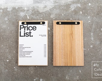 May include: Two rectangular wooden menu boards with black metal clips and gold-toned fasteners. The left board holds a white sheet with the black text Price List, July '23, business hours, and a list of services with prices in US dollars.