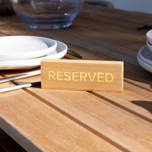 Könnte beinhalten: Ein Holzschild mit dem Wort "RESERVED" in goldfarbenen Buchstaben steht auf einem Holztisch, neben weißen Tellern, Besteck und einer Glasschüssel. Der Tisch ist für eine Mahlzeit gedeckt.