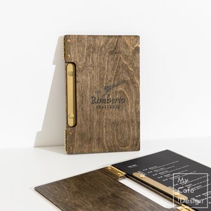 May include: A dark brown wooden menu cover featuring a gold-toned metal spine and hinge mechanism. The front cover is engraved with the text Rimberio TRATTORIA and a small graphic. A second open menu cover with a black insert is visible in the foreground.