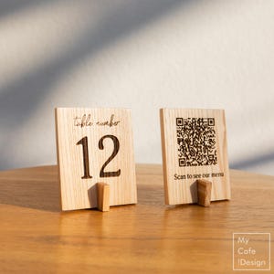 Könnte beinhalten: Zwei hölzerne Tischnummernschilder. Das erste Schild trägt den Text "table number 12" und das zweite Schild trägt einen QR-Code mit dem Text "Scan to see our menu".