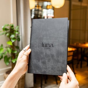 May include: A dark grey menu with the word "luna" embossed on the front. The menu is held in front of a restaurant setting. The restaurant's name is "luna - in the garden". The background includes a plant and a table.