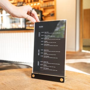May include: A vertical rectangular menu sign with a black background and white text, held in a black base with two gold-colored circular accents. The menu lists food items under categories including Starter, Main Dish, and Dessert in white font.