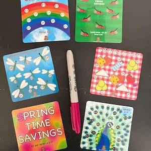 May include: A collection of colorful savings cards with various themes, including a rainbow, lawn mowers, dragonflies, a peacock, and picnic items. A pink Sharpie marker is also present. The cards feature text and illustrations.