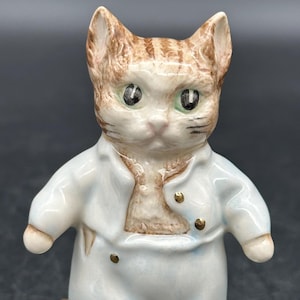 May include: A porcelain figurine of a cat wearing blue overalls and a white shirt. The cat has brown and white fur and green eyes. The figurine is standing on a green base.