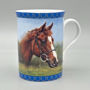 May include: A ceramic mug featuring a detailed illustration of a brown horse's head with a white blaze and bridle. The mug has a blue rim and base with a repeating circle pattern. The handle is white.