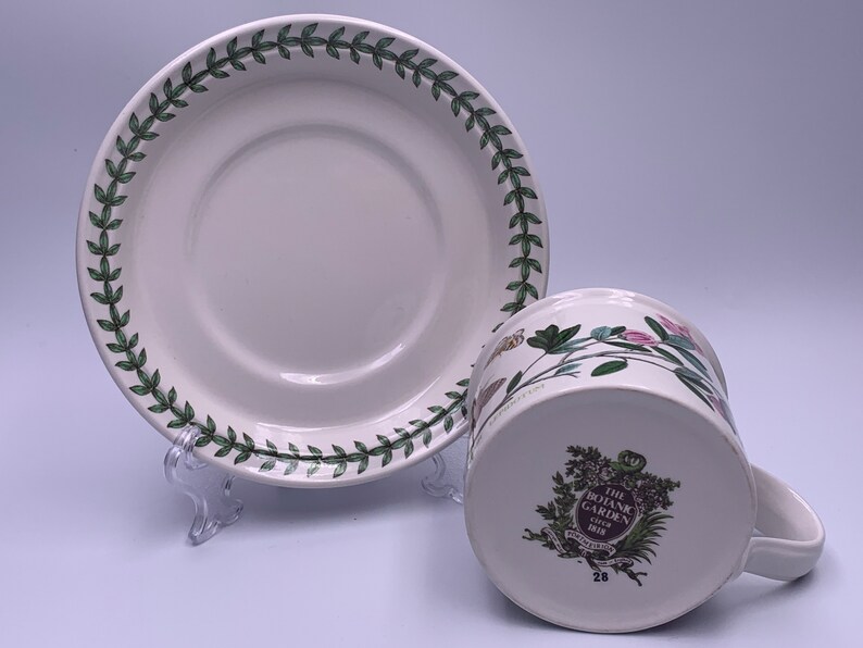 May include: A white teacup and saucer with a green floral pattern. The saucer has a green border with a repeating leaf design. The teacup has a floral design with pink, green, and yellow flowers. The bottom of the teacup has a logo that reads "The Botanic Garden, Giftware, 28".