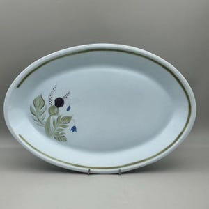 May include: Oval serving platter with a light blue surface and a green border. The platter features a floral design with green leaves, purple berries, and blue bell-shaped flowers. The platter is supported by a metal stand.