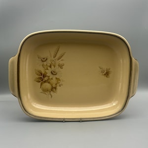 May include: A rectangular, cream-colored serving dish with a floral design. The dish has a slightly raised rim and two handles. The floral design features wheat, flowers, and fruit in shades of brown and tan. The dish is likely made of ceramic or porcelain.