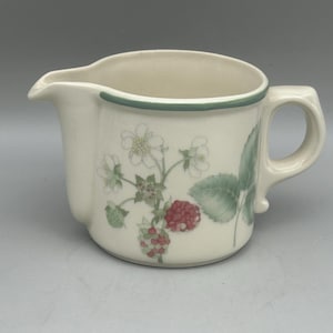 May include: A cream-coloured ceramic creamer jug featuring a botanical illustration of red raspberries, green leaves, and white flowers. The jug has a curved handle, a pouring spout, and a thin green band painted along the top rim.