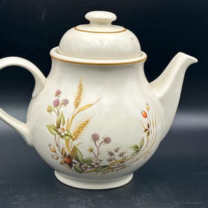 May include: Cream-colored ceramic teapot with a lid, handle, and spout. The teapot is decorated with a floral and wheat design in shades of brown, green, and red. The rim and lid are trimmed with a gold-colored band.