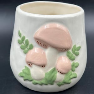 Può includere: Tazza in ceramica bianca con un design di funghi rosa e bianchi. La tazza ha foglie verdi e steli marroni sui funghi.