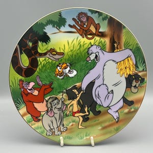 Può includere: Un piatto decorativo con personaggi de Il libro della giungla di Disney. Il piatto mostra Baloo, Bagheera, Mowgli e altri personaggi in una colorata scena della giungla. Il piatto ha un bordo dorato ed è esposto su un supporto bianco.