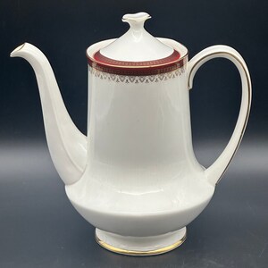 Peut inclure: Cafetière en porcelaine blanche avec une bande décorative rouge et dorée autour du couvercle. La poignée et le bec sont blancs avec des bordures dorées. Le pot a un bec courbé et une base arrondie.