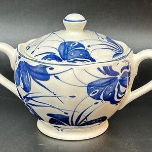 May include: A white ceramic sugar bowl with a lid, featuring a blue and white floral design. The bowl has a rounded shape and two handles.