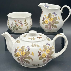 Könnte beinhalten: Ein weißes Tee-Set bestehend aus Teekanne, Zuckerdose und Milchkännchen mit einem Blumenmuster mit Primeln und Schmetterlingen. Das Set ist mit dem Text "Primula Vulgaris" verziert.