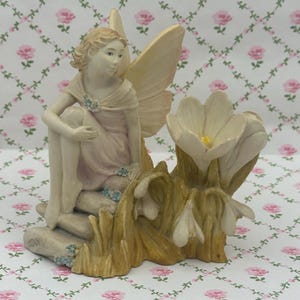 May include: A fairy figurine with cream-coloured wings and a pale pink dress, seated beside white flowers with yellow centres. The base of the figurine has a stone-like appearance with small blue flower accents. The figurine is a decorative item.