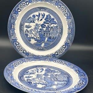 May include: Two blue and white ceramic dinner plates with a traditional willow pattern. The plates feature a blue and white design of a willow tree, a pagoda, a bridge, and a boat.