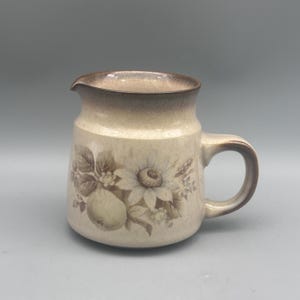 May include: A ceramic creamer pitcher featuring a speckled cream-colored glaze with a brown rim and handle. The body is decorated with a muted brown botanical illustration of a large sunflower, leaves, and a round fruit, creating a rustic appearance.