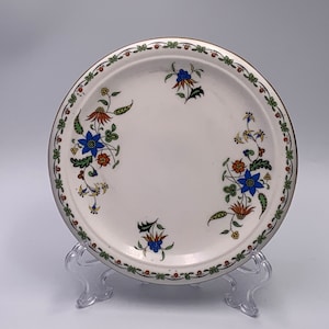 May include: A white ceramic plate with a floral design in shades of blue, green, orange, and yellow. The plate has a gold rim and is decorated with a repeating pattern of flowers and leaves.