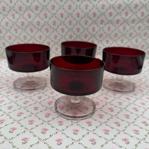 May include: Four vintage dessert glasses. Each glass has a deep red bowl and a clear stem. The glasses are displayed on a floral patterned surface. The bowls have a textured appearance.