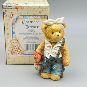 May include: A tan teddy bear figurine wearing a white head wrap, a white vest, and blue trousers, wrapped in a dark chain and holding a small beige bag with a dollar sign. The bear is positioned in front of its original cardboard box packaging.
