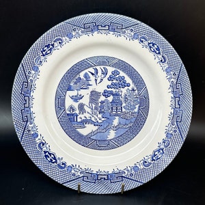 May include: A circular, white ceramic plate with a blue Willow pattern. The plate showcases a central scene with a pagoda, bridge, and birds, framed by a detailed border. The plate is presented on a black surface.