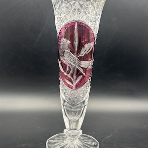 May include: A clear crystal vase with a flared top and a circular base. The vase features a detailed bird and foliage design in a deep magenta oval. The vase has a cut crystal pattern and is approximately 25 cm tall.