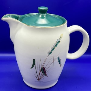 Peut inclure: Une théière en céramique blanche avec un couvercle vert et un motif floral composé de feuilles et de tiges vertes, rouges et blanches.