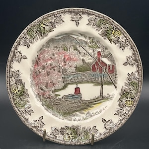 May include: A vintage ceramic plate with a brown and green floral border. The centre of the plate features a colourful illustration of a country scene with a red house, trees, and a pond.