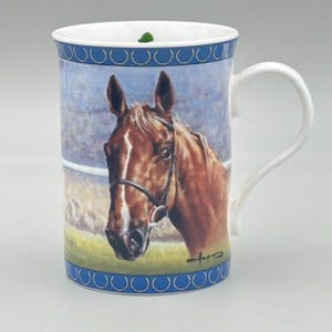 May include: White ceramic mug featuring a detailed portrait of a brown horse with a black bridle. The mug has a blue border with a repeating pattern and a white handle. The background shows a blurred image of a race track.