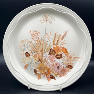 Peut inclure: Une assiette plate en céramique blanche avec un motif floral représentant du blé, des fleurs sauvages et des feuilles dans des tons de brun, orange et bleu.
