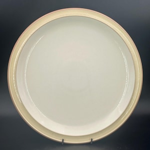 May include: A round, off-white ceramic plate with a light tan and pink rim. The plate is a simple design, suitable for everyday use. The plate is displayed on a black surface.