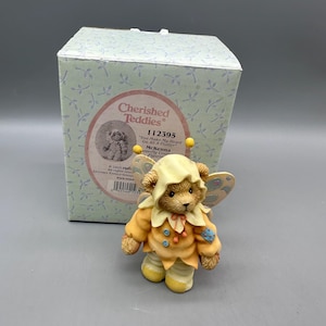 May include: A Cherished Teddies figurine named McKenna, featuring a bear in a yellow butterfly costume with wings, standing before its original light blue box with a leaf pattern. The box displays the text Cherished Teddies 112395 You Make My Heart Go All A Flutter.